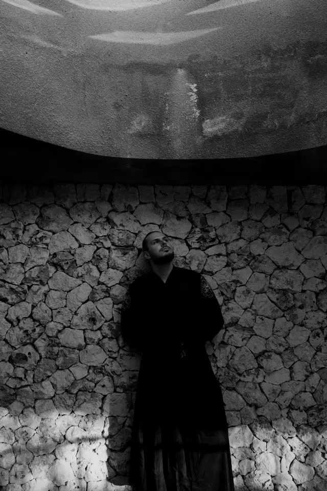 OPPAACHA standing against a rough stone wall under a concrete canopy, black and white